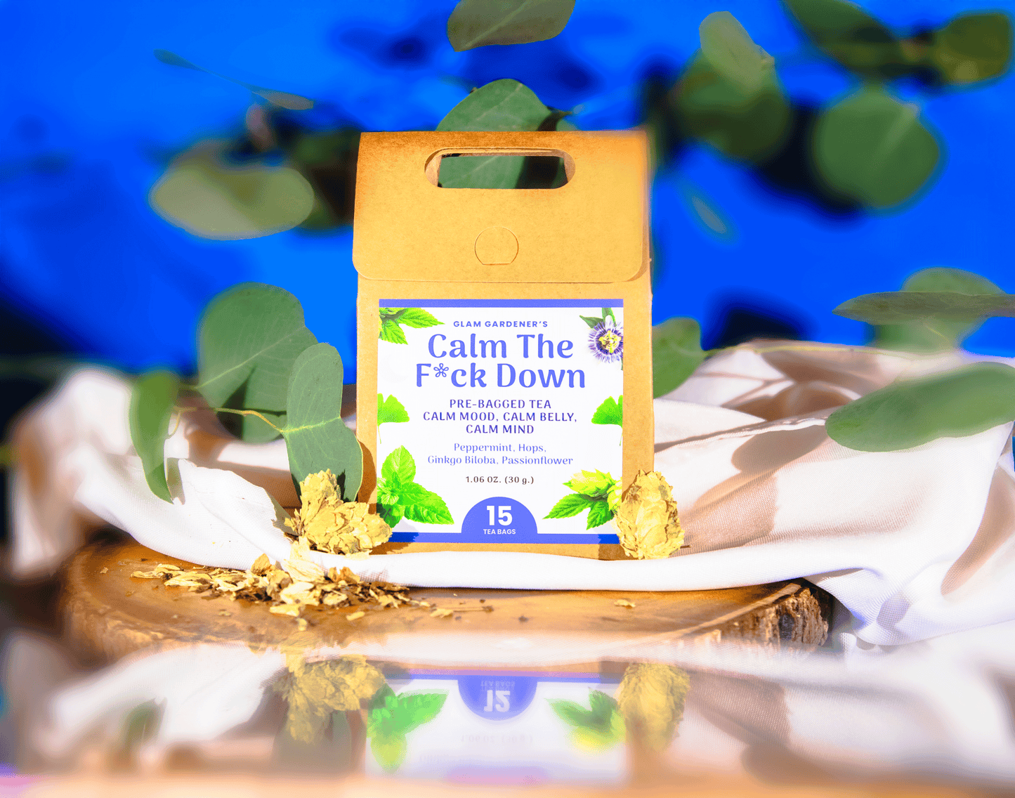 A product package for 'Calm The F*ck Down' peppermint tea by Slow Garden's sits on a white cloth and wooden slice, surrounded by green leaves and loose tea leaves against a vibrant blue background. Hops, Peppermint, Ginkgo Biloba, Passionflower