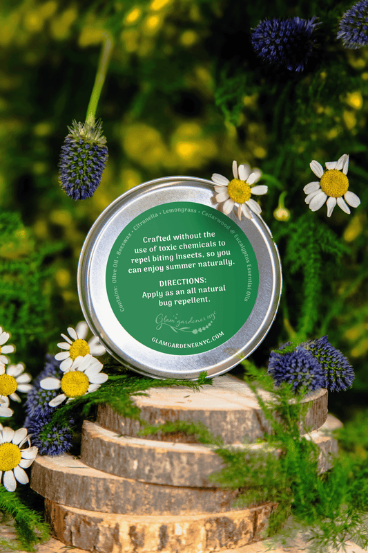 A photo of Glam Gardener NYC’s Bug Balm Natural Bug Repellent featuring a floral background. The product is in a 2 ounce aluminum tin that features a screw cap. It is all natural and crafted with beeswax, olive oil, and essential oils including citronella, eucalyptus, cedarwood, and lemongrass. This is showcasing the back label.