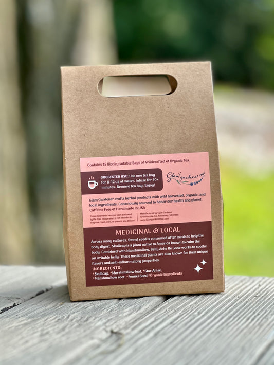 A kraft paper reusable tea bag bag with a die-cut handle stands upright on a weathered gray wooden surface outdoors. The bag has a pink and dark brown printed label with text and a logo that appears to read Glam Gardener NYC. Across many cultures, fennel seed is consumed after meals to help the body digest. Skullcap is a plant native to America known to calm the body. Combined with Marshmallow, Belly Ache Be Gone works to soothe an irritable belly.
