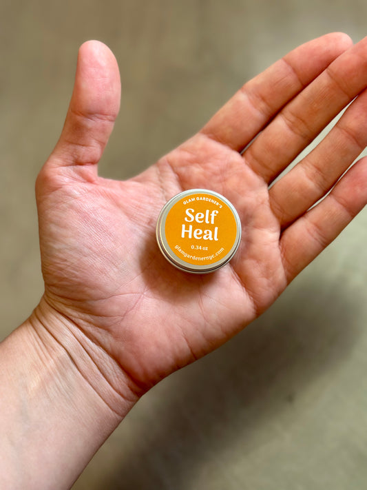 Glam Gardeners Self Heal Mini in hand to show the small size of the mini salve. The yellow container labeled Glam Gardener's Self Heal sits in the palm of someone's hand.