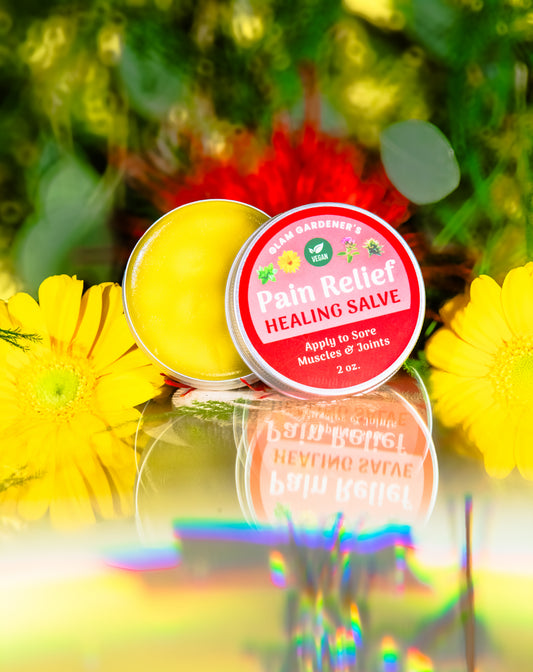 Glam Gardener's Vegan Pain Relief is a healing salve in a 2 ounce tin with a red label. The vegan emblem is centered on the label that also reads "Glam Gardener's Pain Relief Healing Salve Apply to Sore Muscles & Joints 2 oz." The background features green, red, and yellow plants. There is a rainbow filter on the image toward the bottom that mirrors a reflection of the salve.