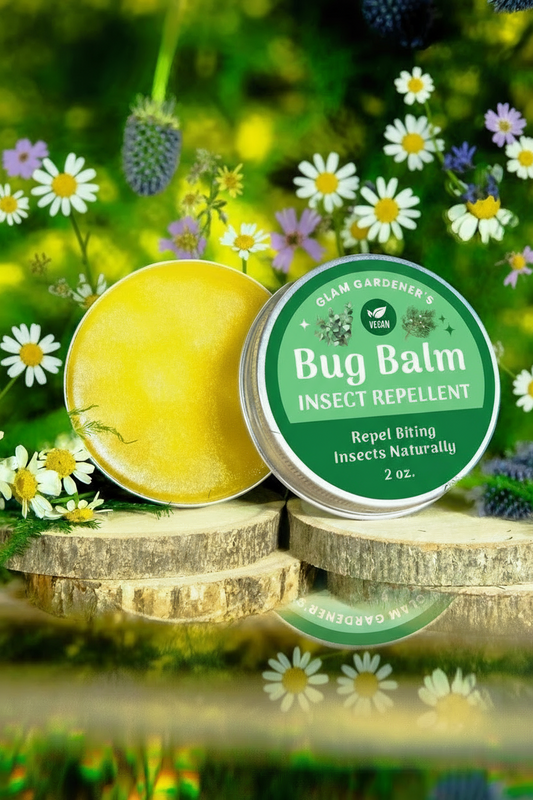 Glam Gardener NYC’s vegan bug balm featuring a 2 ounce aluminum tin with the balm inside of the tin. This is a 100% plant based formula with olive oil, candle wax, and essential oils, including eucalyptus, lemongrass, citronella, and Cedarwood. The background of this image features a floral and forest like background. This product is an all natural alternative for bug repellent designed to be applied on the skin.
