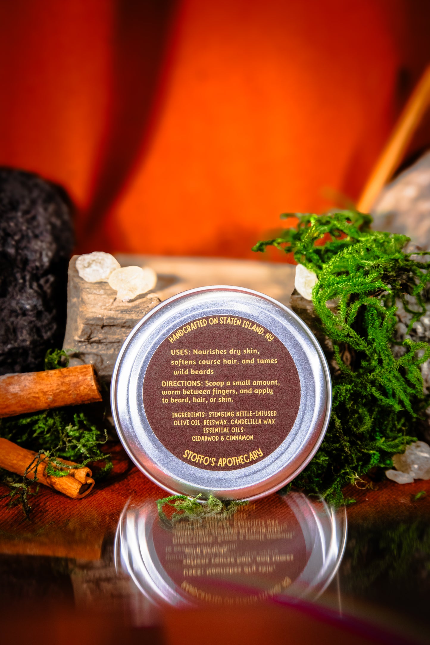 A round metal tin of Stoffo's Apothecary Glam Gardener Beard Balms hair and scalp massage oil, styled on a reflective dark surface with moss, crystals, cinnamon sticks, and a warm orange background.