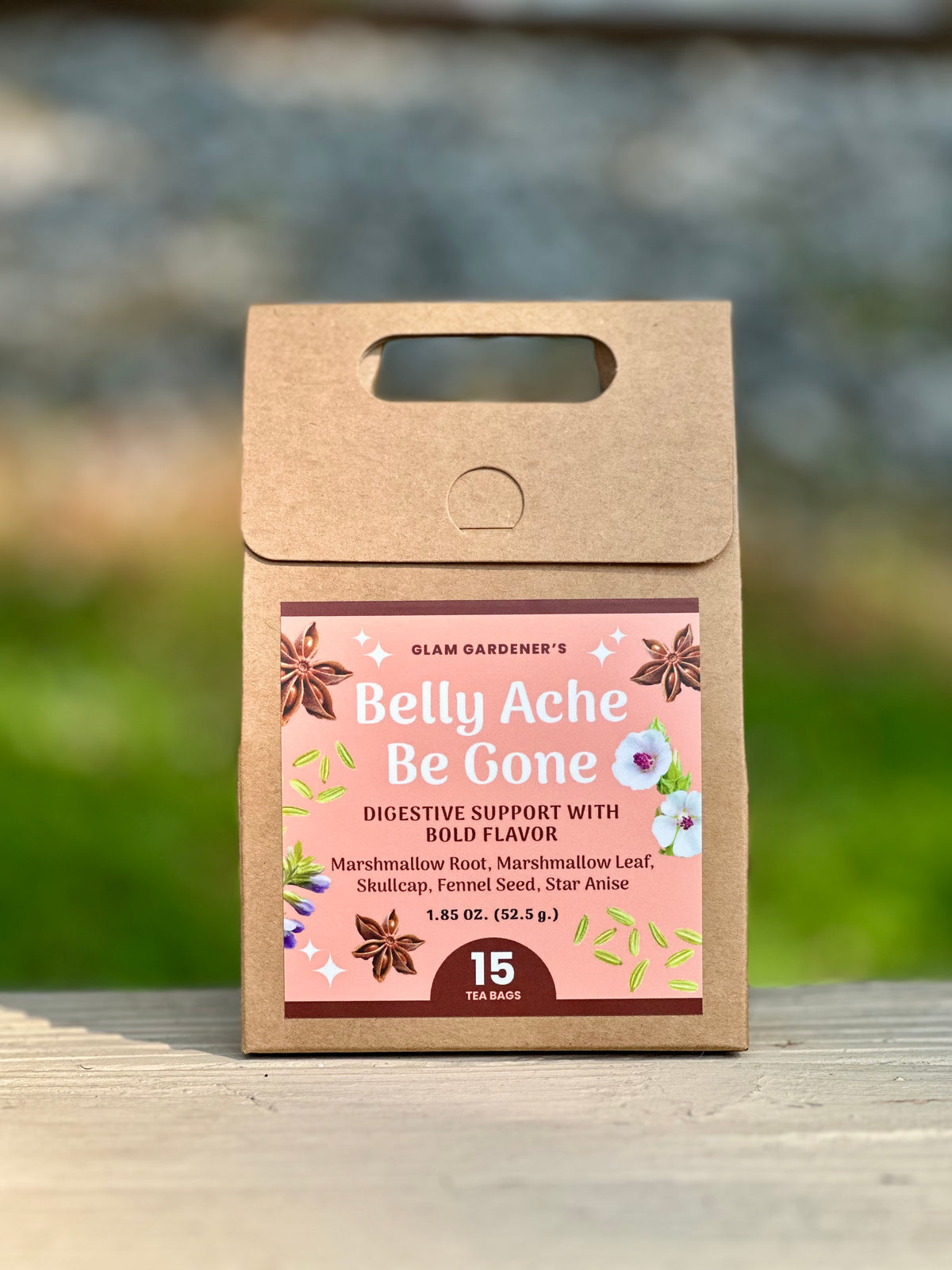 A kraft paper tea bag pouch labeled 'Glam Gardener's Belly Ache Be Gone' stands upright on a wooden surface with a blurred outdoor background. The ingredients read Marshmallow leaf, Marshmallow root, Star Anise, Fennel Seed and Skullcap