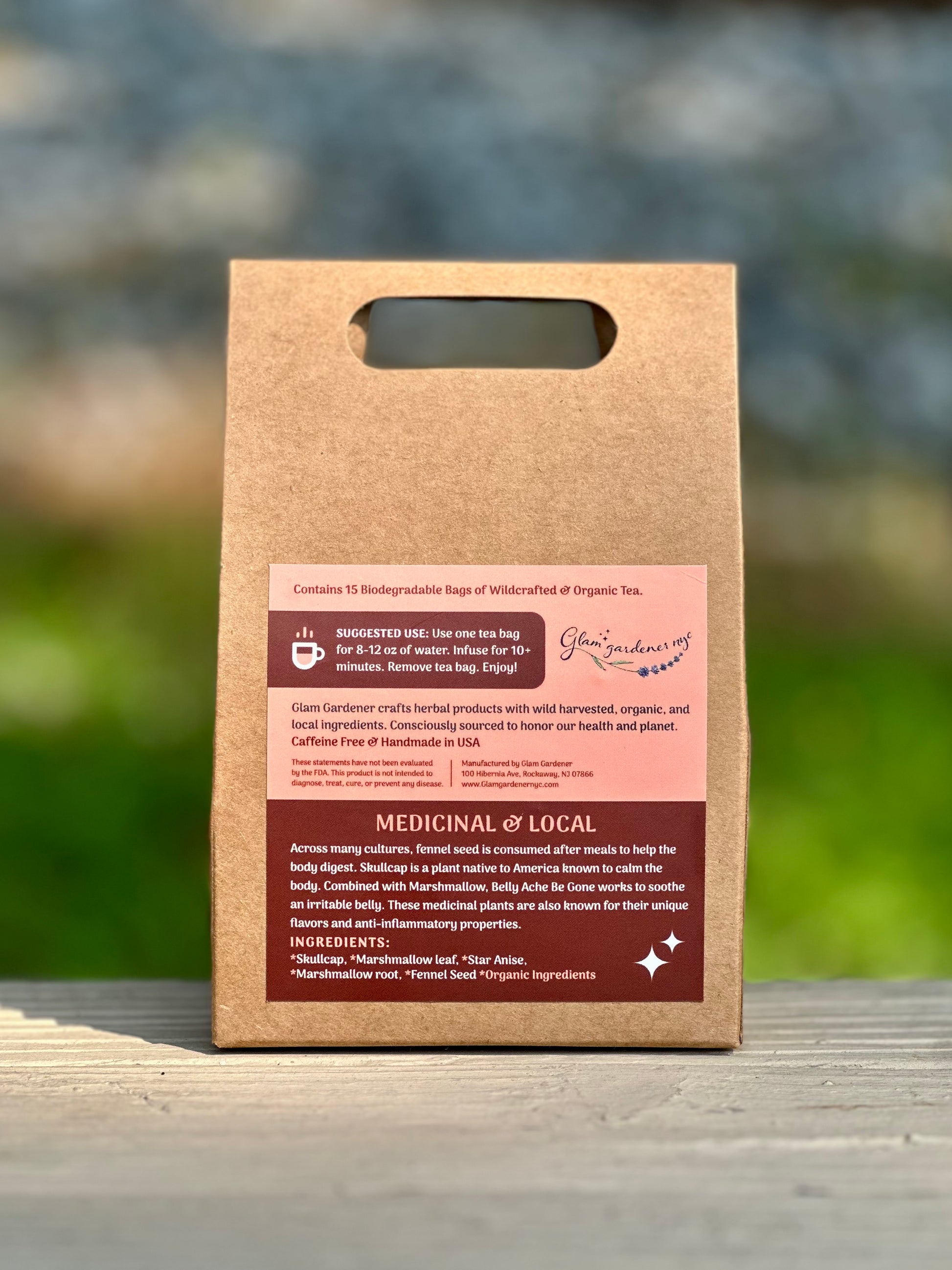 A kraft paper tea bag pouch with a handle stands upright on a wooden surface outdoors. The front label is mostly white with dark brown and pink sections, reading 'Medicinal & Local' and 'Caffeine Free. Across many cultures, fennel seed is consumed after meals to help the body digest. Skullcap is a plant native to America known to calm the body. Combined with Marshmallow, Belly Ache Be Gone works to soothe an irritable belly. These medicinal plants are also known for their unique flavors and anti-inflammator