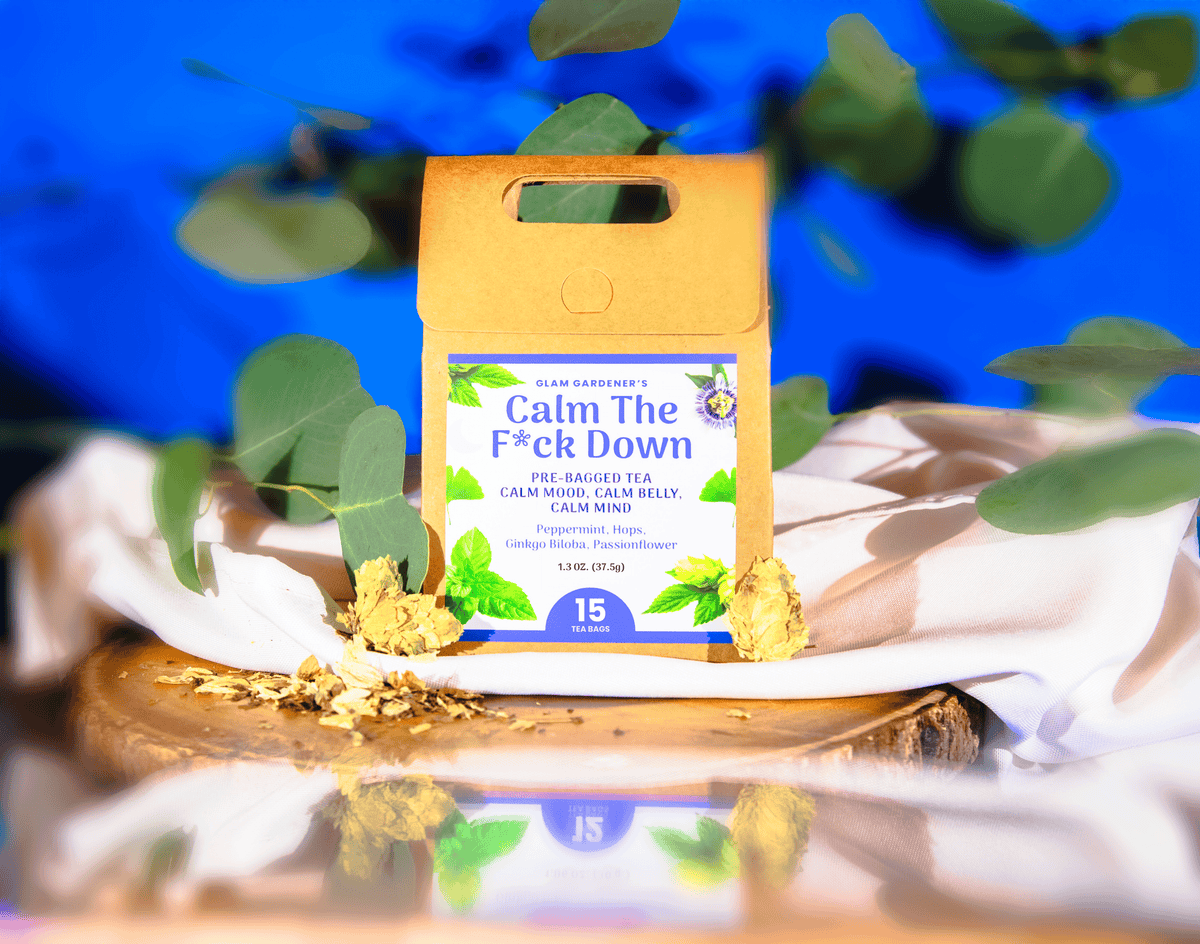 A product package for 'Calm The F*ck Down' peppermint tea by Slow Garden's sits on a white cloth and wooden slice, surrounded by green leaves and loose tea leaves against a vibrant blue background. Hops, Peppermint, Ginkgo Biloba, Passionflower