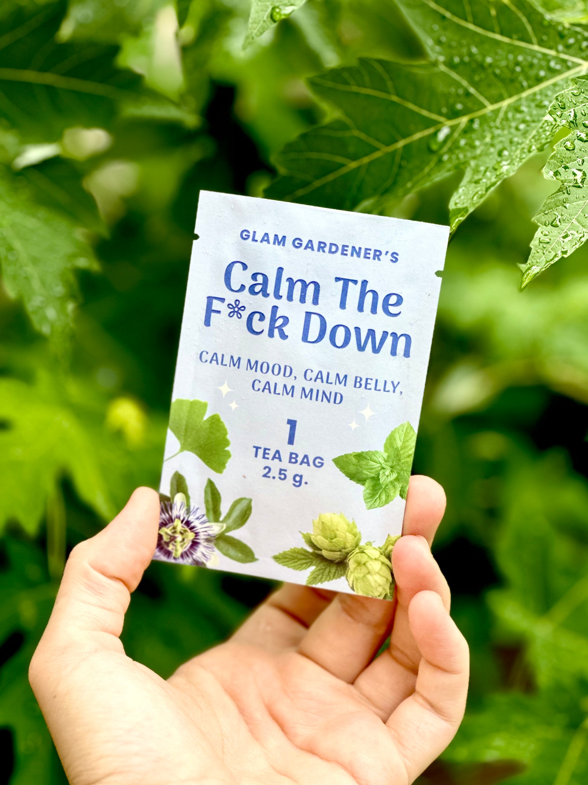 A hand holds a single tea bag packet labeled 'Glam Gardener's Calm The F*ck Down' against a background of green garden leaves.