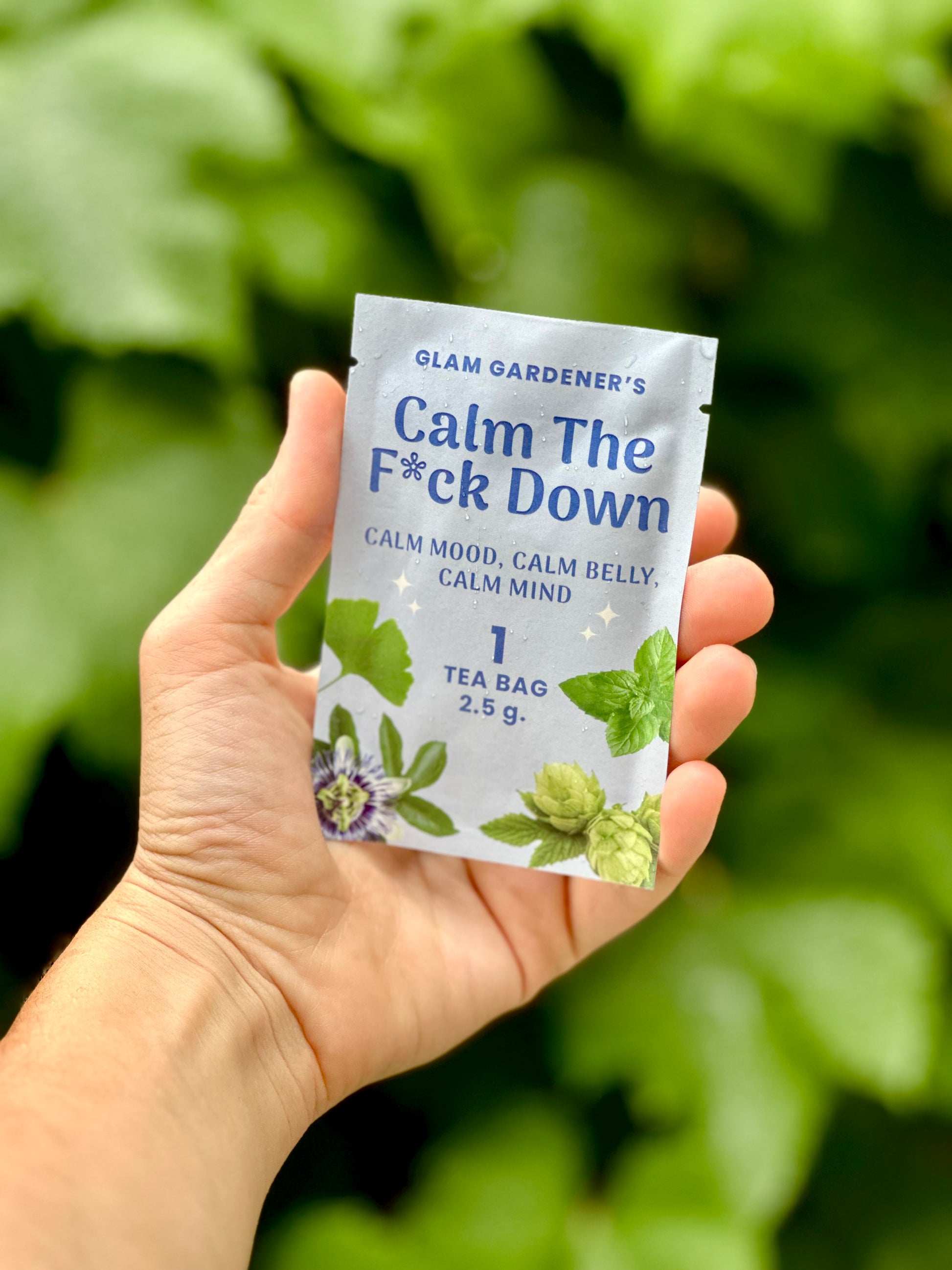 A hand holding a single tea bag packet labeled 'Glam Gardener's Calm The F*ck Down' against a blurred green leafy background.