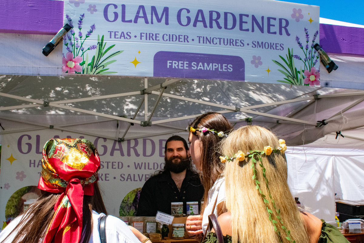 Glam Gardener is a business based in New York with a commercial kitchen in New Jersey that handcrafts all items that we sell! We make herbal tea, fire cider, tinctures, and more. Glam Gardener attends markets in New Jersey, New York, and beyond selling and sampling herbal products. This is a picture of Glam Gardener's booth taken at the New Jersey Renaissance Faire. The image shows workers at the Glam Gardener booth and customers enjoying their offerings. The booth is white and purple and the sky is blue.