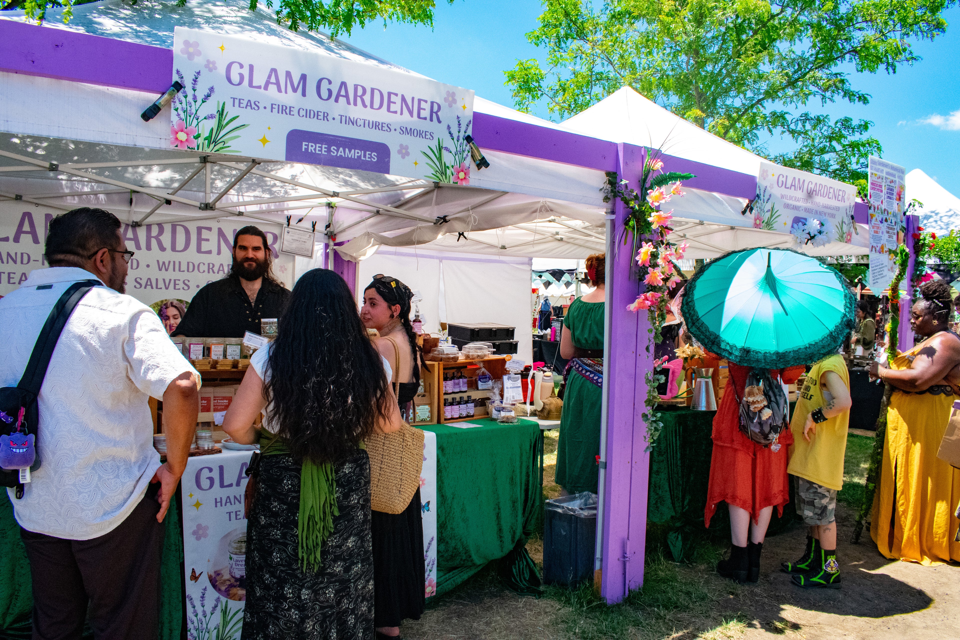 Glam Gardener is a business based in New York with a commercial kitchen in New Jersey that handcrafts all items that we sell! We make herbal tea, fire cider, tinctures, and more. Glam Gardener attends markets in New Jersey, New York, and beyond selling and sampling herbal products. This is a picture of Glam Gardener's booth taken at the New Jersey Renaissance Faire. The image shows workers at the Glam Gardener booth and customers enjoying their offerings. The booth is white and purple and the sky is blue.