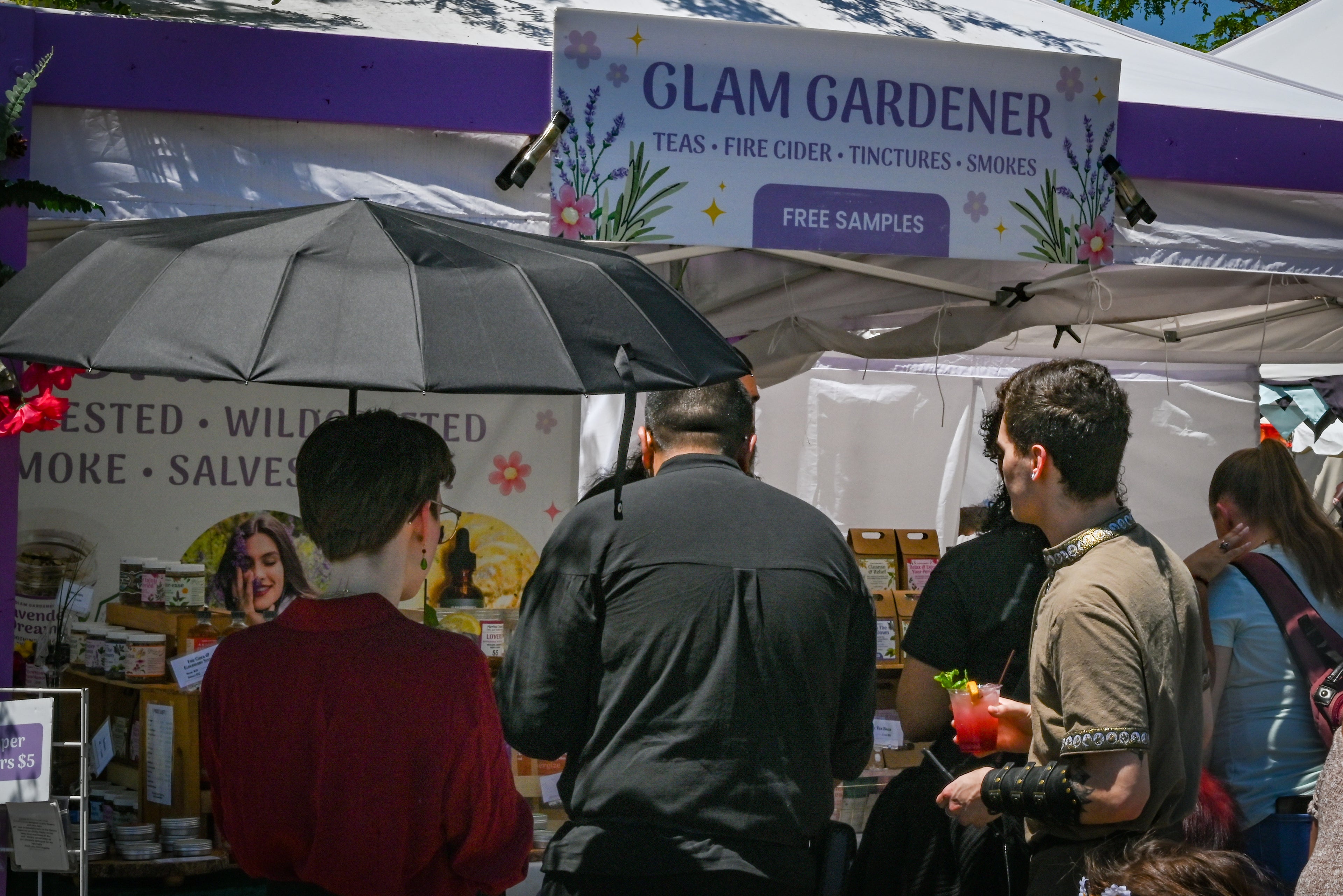 Glam Gardener is a business based in New York with a commercial kitchen in New Jersey that handcrafts all items that we sell! We make herbal tea, fire cider, tinctures, and more. Glam Gardener attends markets in New Jersey, New York, and beyond selling and sampling herbal products. This is a picture of Glam Gardener's booth taken at the New Jersey Renaissance Faire. The image shows workers at the Glam Gardener booth and customers enjoying their offerings. The booth is white and purple and the sky is blue.