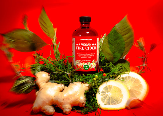 Glam Gardener’s Spicy Fire Cider. The image shows an 8 ounce glass bottle of the fire cider on a red backdrop surrounded by plants that are in the infusion. The infusion includes apple cider vinegar, raw and organic, honey, ginger, turmeric, spicy peppers, wild spicebush and more.