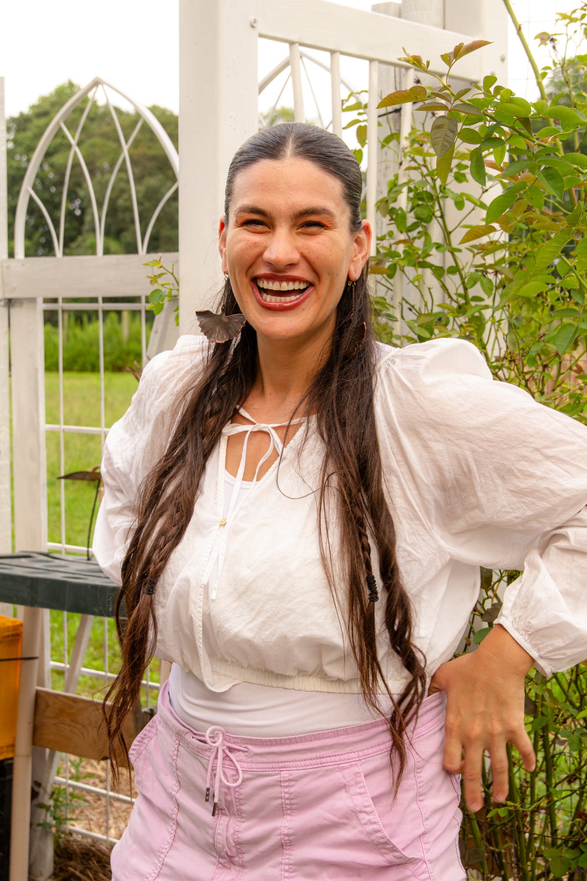 A woman named Aly Stoffo, Owner of Glam Gardener NYC, with long dark hair wearing a white long-sleeve blouse with a tie neckline and light pink drawstring shorts, standing outdoors in front of a white garden trellis and greenery.