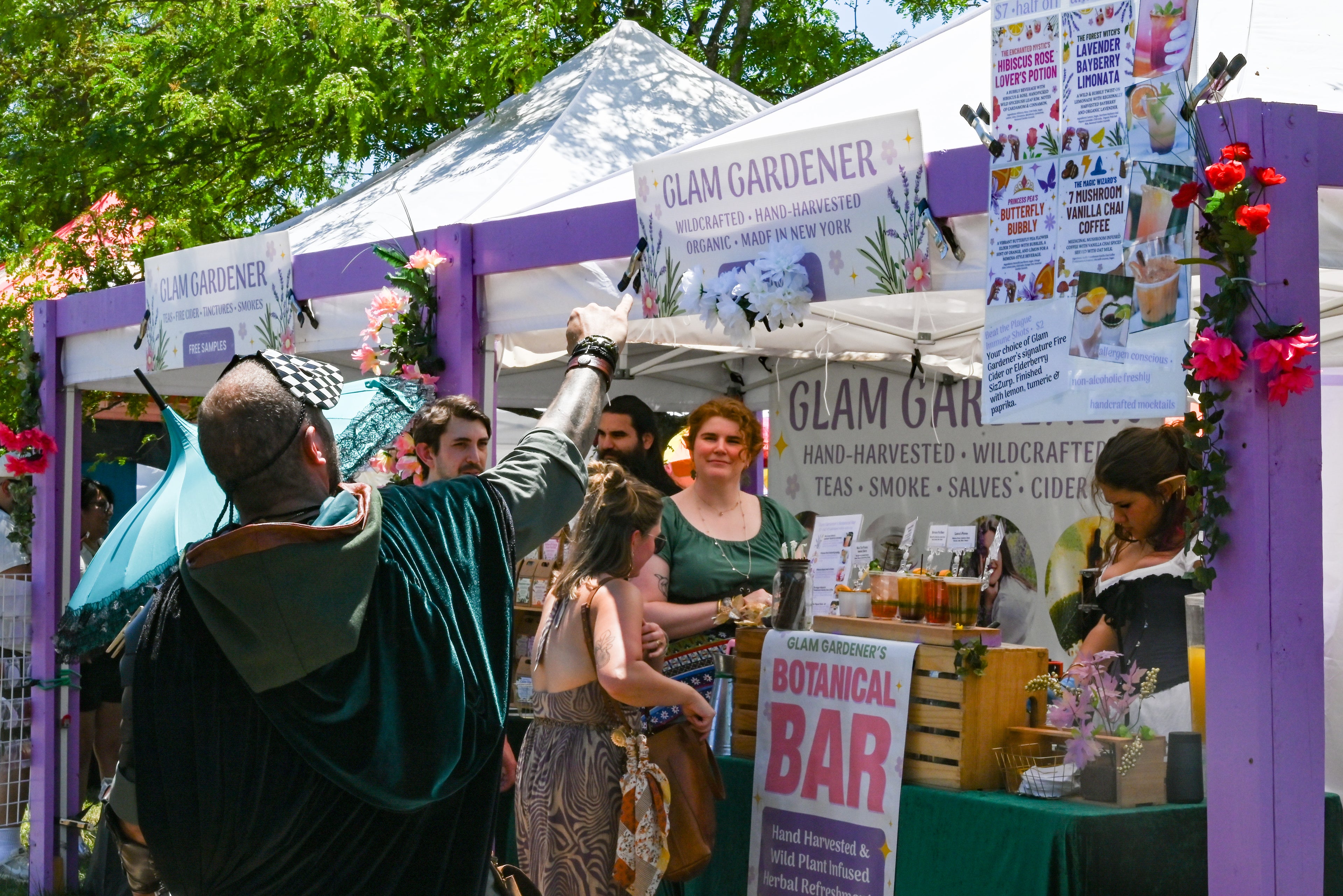 Glam Gardener is a business based in New York with a commercial kitchen in New Jersey that handcrafts all items that we sell! We make herbal tea, fire cider, tinctures, and more. Glam Gardener attends markets in New Jersey, New York, and beyond selling and sampling herbal products. This is a picture of Glam Gardener's booth taken at the New Jersey Renaissance Faire. The image shows workers at the Glam Gardener booth and customers enjoying their offerings. The booth is white and purple and the sky is blue.