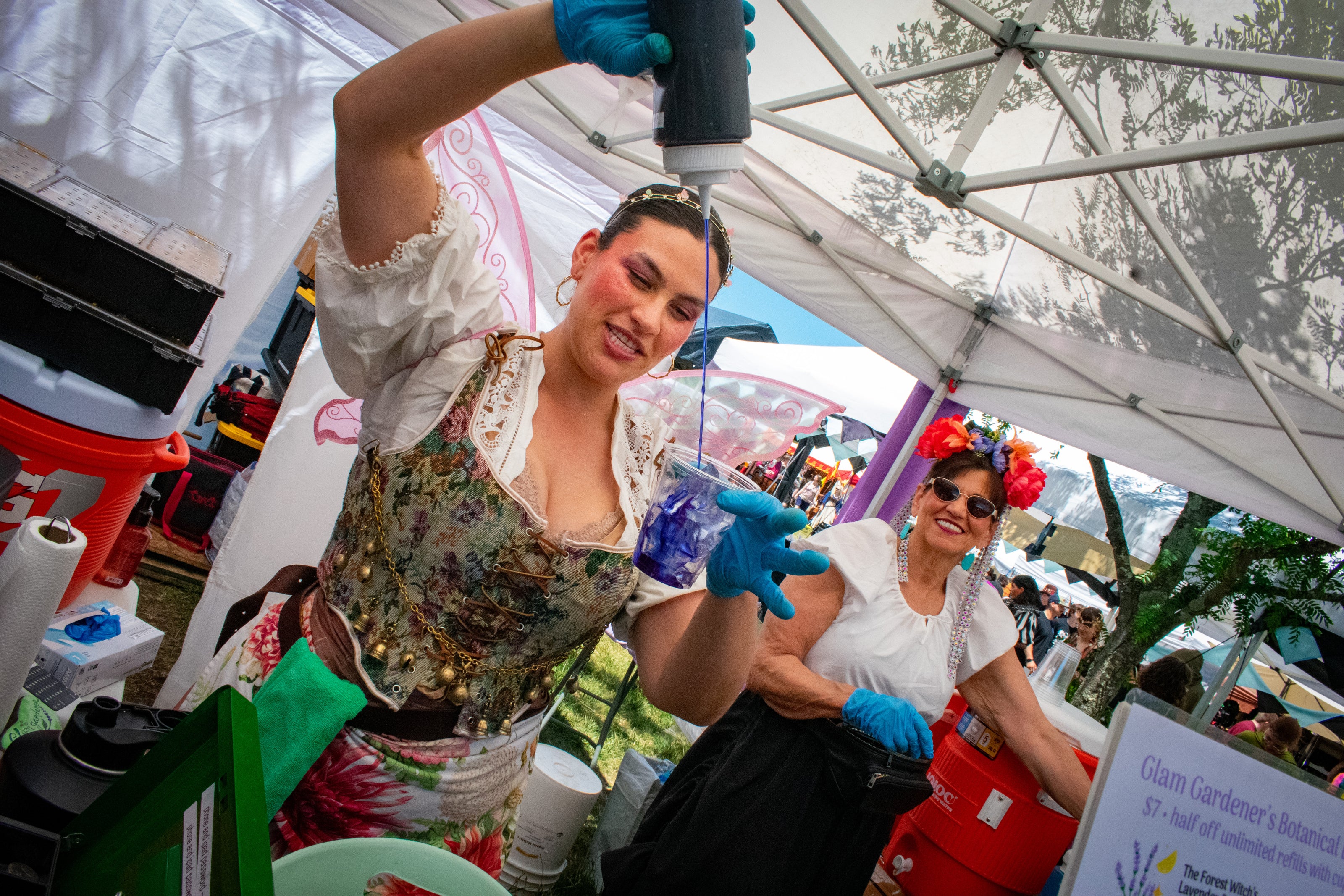 The Glam Gardener serving her signature Botanical Bar beverages at the New Jersey Renaissance Faire. The Glam Gardener is an herbal potion creator that combines the benefits of medicinal plants with signature non-alcoholic beverages. Creating fantastical experiences for enjoyers of the Botanical Bar across NYC, New Jersey, and beyond! The Glam Gardener is wearing fairy wings and a corset at the New Jersey Renaissance Faire. The beverage she is pouring is blue. Glam Gardener's Botanical Bar is for hire.