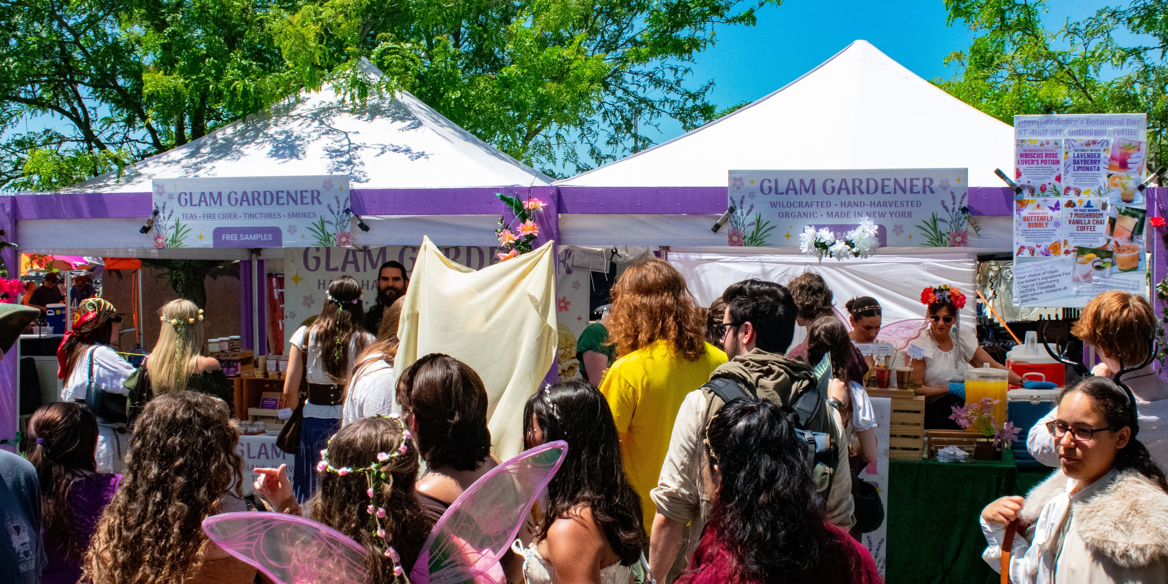 Glam Gardener NYC and Glam Gardener's Botanical Bar at the New Jersey Renaissance Faire. This image shows a crowd of customers dressed in cosplay lining up in front of Glam Gardener's apothecary booth that sells herbal tea, fire cider, tinctures, wildcrafted and handcrafted items made in New York. The booth is a lavender colored purple and it's providing structural support to 2 white canopies. There is a sunny sky that is blue in the background as well as trees that exist on the site of the fairgrounds.