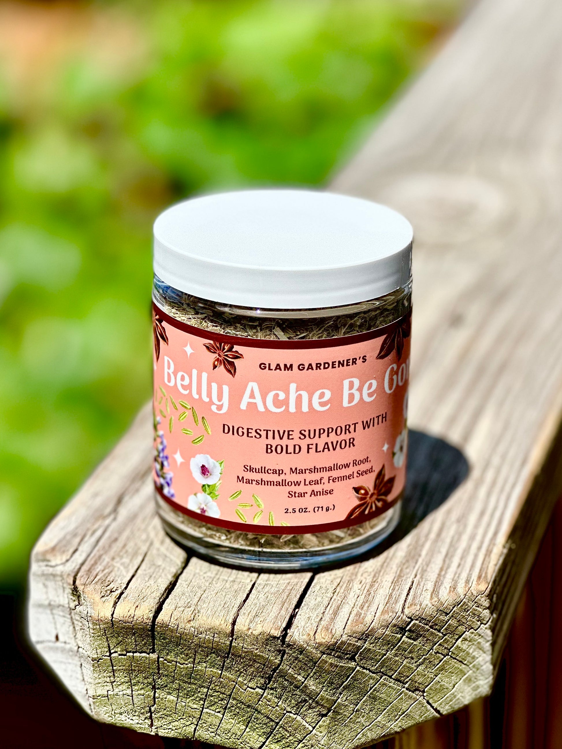 A jar of Glam Gardener's Belly Ache Be Gone digestive support supplement, featuring a pink label and white lid, resting on a weathered wooden plank with a blurred green garden background.