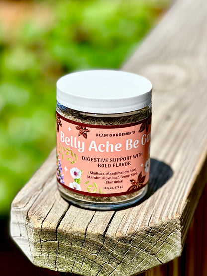 A jar of Glam Gardener's Belly Ache Be Gone digestive support supplement, featuring a pink label and white lid, resting on a weathered wooden plank with a blurred green garden background.