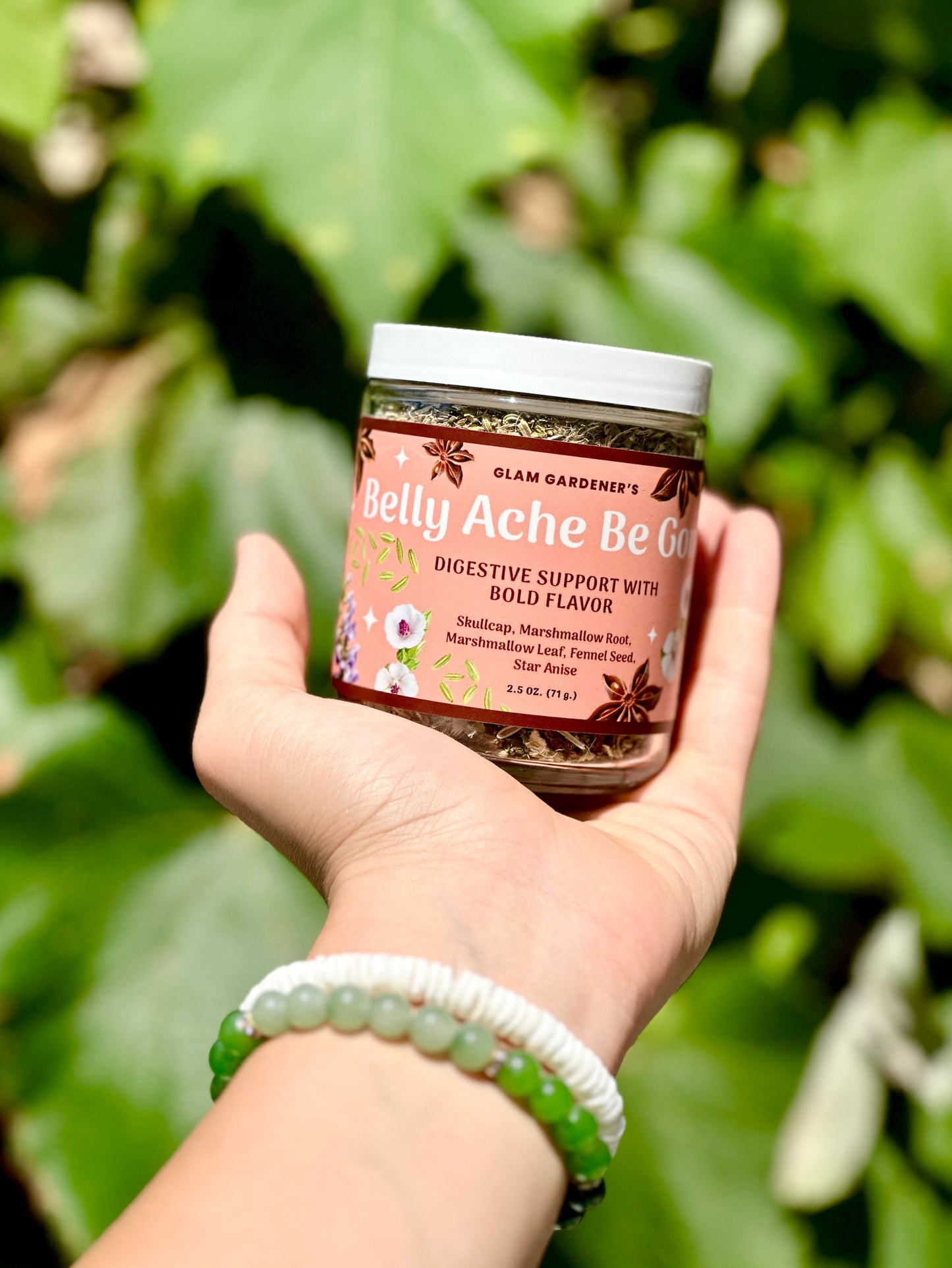A hand holding a jar of Glam Gardener's Belly Ache Be Gone digestive support supplement, set against a blurred green garden background.