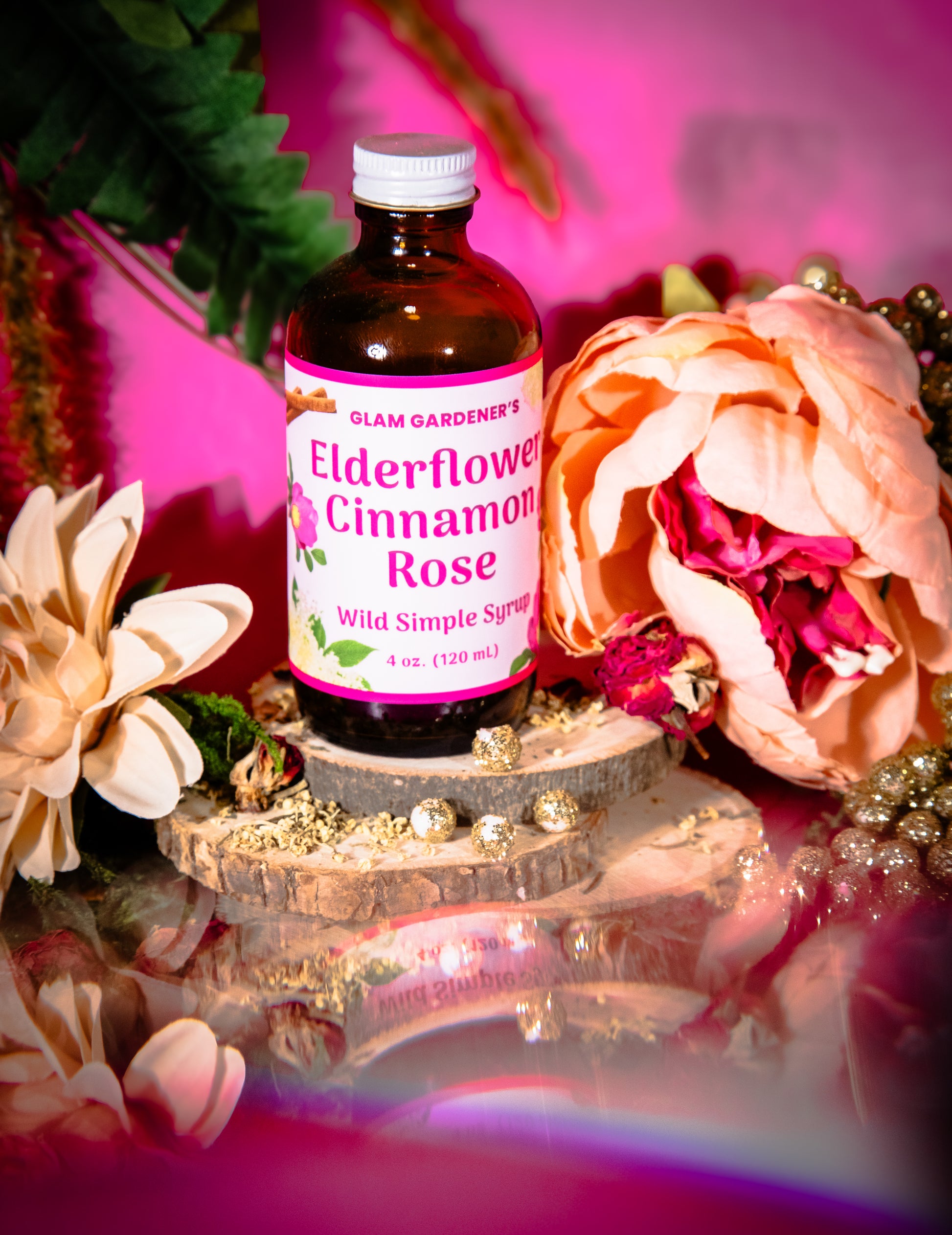 A bottle of Glam Gardener's Elderflower Cinnamon Rose Wild Simple Syrup displayed on a decorative wooden slice pedestal. The amber glass bottle has a white cap and a pink label with green and white text.
