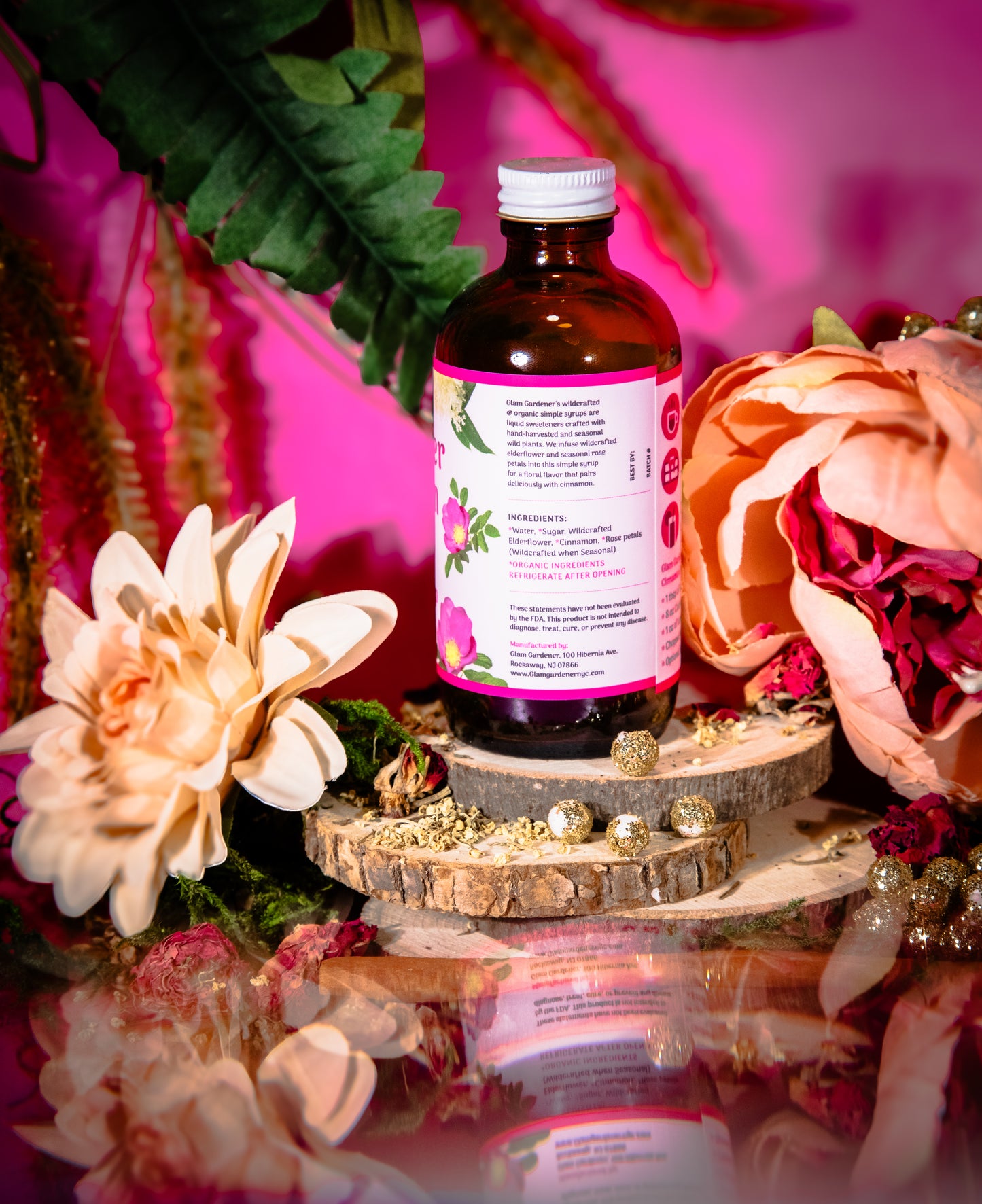 An amber glass bottle with a white cap and a pink label featuring floral illustrations, displayed on a rustic wood slice pedestal surrounded by dried flowers, green leaves, and pink background elements.