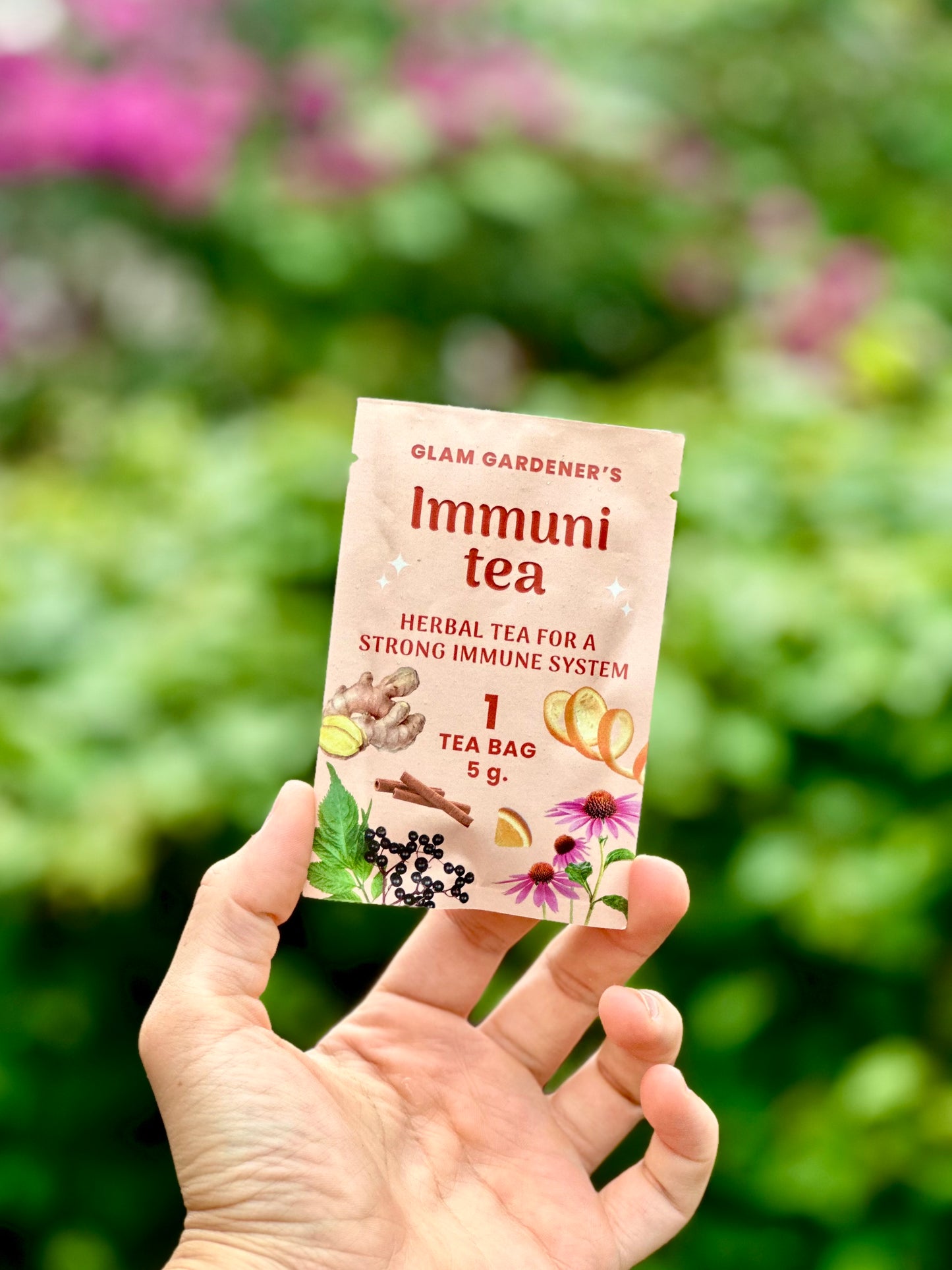 A hand holds a single tea bag packet labeled 'Glam Gardener's Immuni tea' against a blurred green garden background with a pink coneflower flower above.