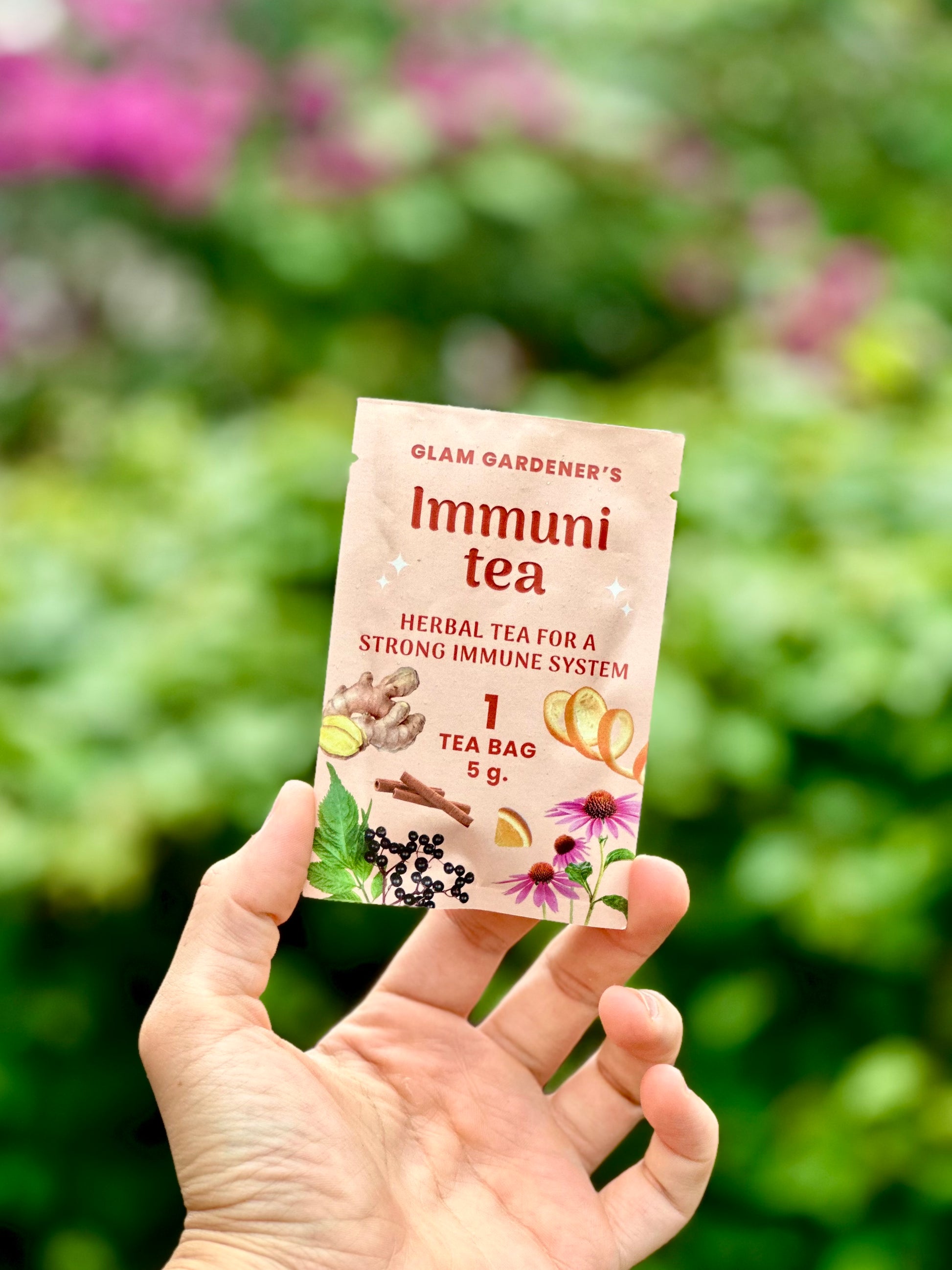 A hand holds a single tea bag packet labeled 'Glam Gardener's Immuni tea' against a blurred green garden background with a pink coneflower flower above.