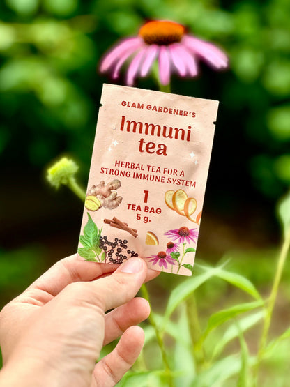 A hand holds a single tea bag packet labeled 'Glam Gardener's Immuni tea' against a blurred green garden background with a pink coneflower flower above.