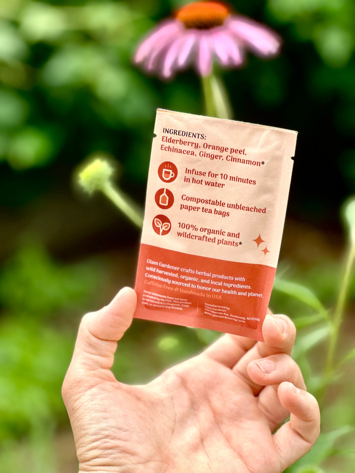 A hand holds a small paper tea bag packet outdoors with a blurred green garden and a pink flower in the background. The packet is cream and terracotta orange with printed ingredient and usage text.