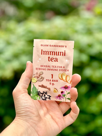 A hand holding a single packet of Glam Gardener's Immuni tea against a blurred green outdoor background.