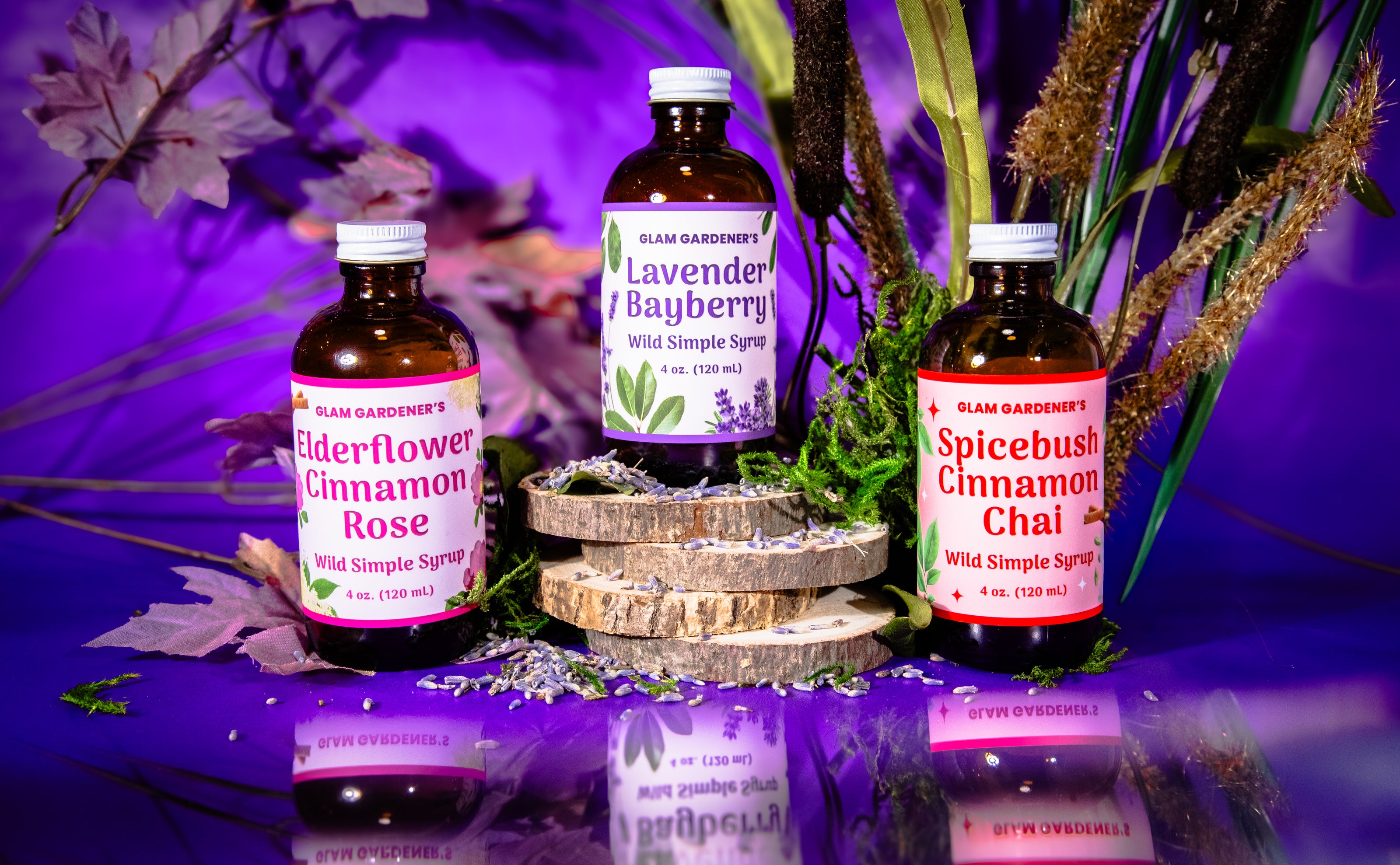 Glam Gardener’s wildflower syrups are mocktail and cocktail crafting syrups made with Wild and regional plants. The photo features three separate wildflower syrups that Glam Gardener offers, elderflower cinnamon rose, spicebush, cinnamon, chai, and lavender Bayberry simple syrup. The photo features three of the 4 ounce bottles on a purple backdrop