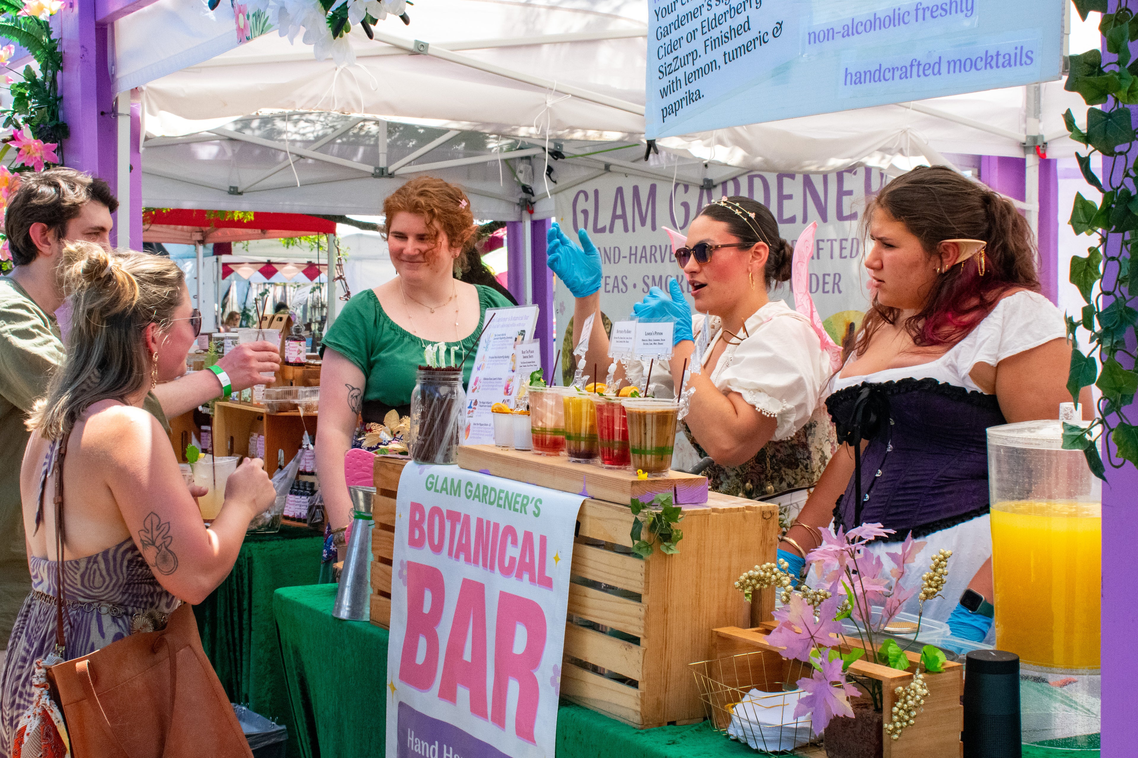 Glam Gardener Serving Customers at the New Jersey Renaissance Faire at the Botanical Bar. A seasonal and handcrafted herbal mocktail service. Glam Gardener's team are dressed like fairies and elves, with pointed ears and fairy wings. 