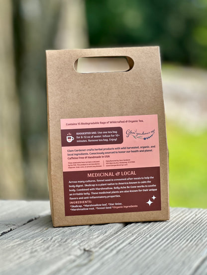 A kraft paper reusable tea bag bag with a die-cut handle stands upright on a weathered gray wooden surface outdoors. The bag has a pink and dark brown printed label with text and a logo that appears to read Glam Gardener NYC. Across many cultures, fennel seed is consumed after meals to help the body digest. Skullcap is a plant native to America known to calm the body. Combined with Marshmallow, Belly Ache Be Gone works to soothe an irritable belly.