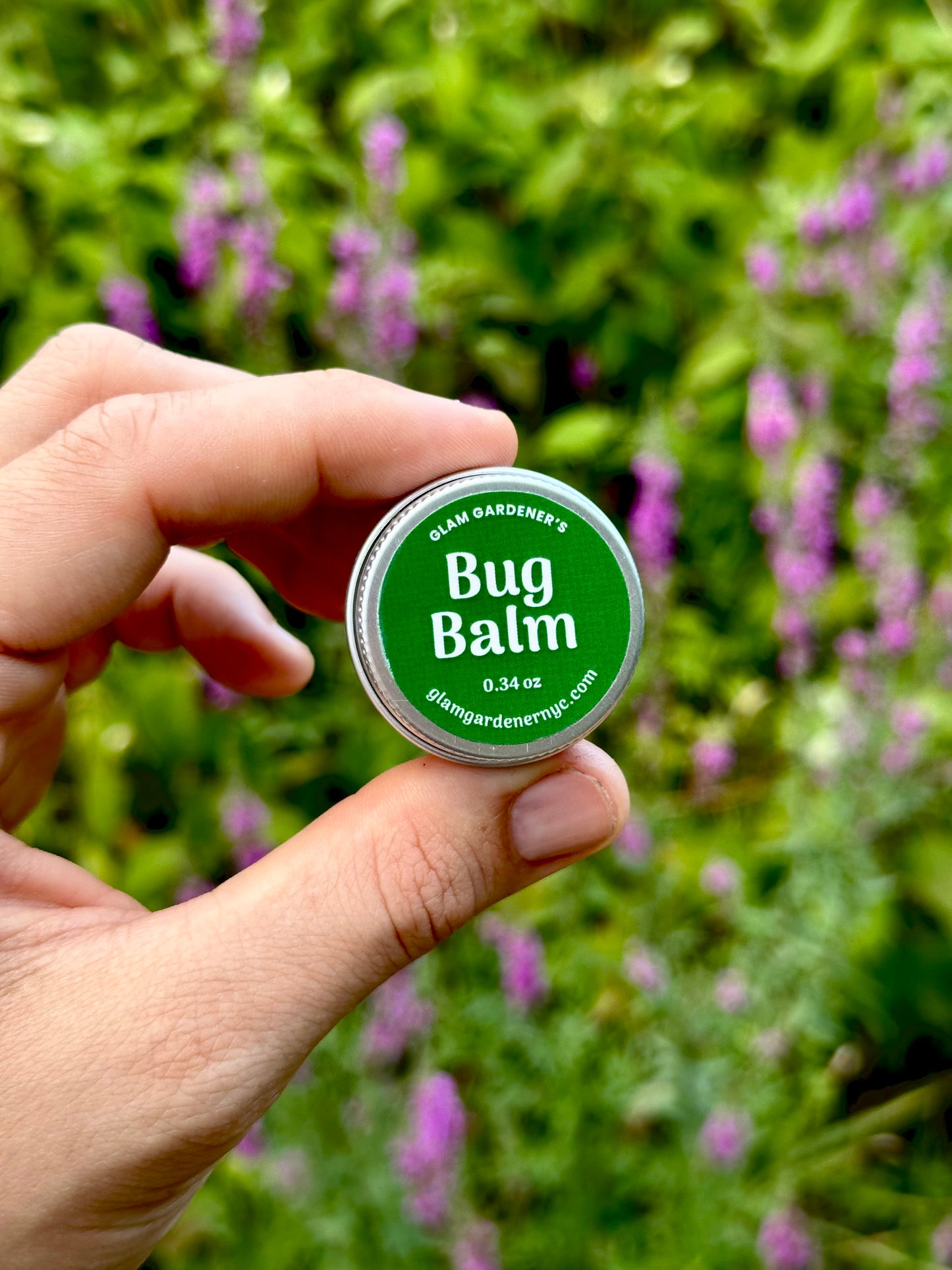 A hand holds a small round tin of Glam Gardener's Bug Balm in front of a blurred garden background with green foliage and purple flowers. This is a natural bug repelling lotion handcrafted and made with essential oils citronella eucalyptus cedarwood and lemongrass