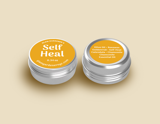 A miniature container of Glam Gardener's Signature Self Heal salve. An herbal salve for cuts, burns and wounds infused with goldenrod, self heal (prunella vulgaris), calendula, and chamomile. The salve is 0.34 ounces and is a more affordable and smaller alternative to their signature Self Heal salve. The image itself shows the aluminum tin that the Self Heal is in. The yellow label has white text that reads on front "Glam Gardener's Self Heal 0.34 oz glamgardenernyc.com. The background is a light yellow.