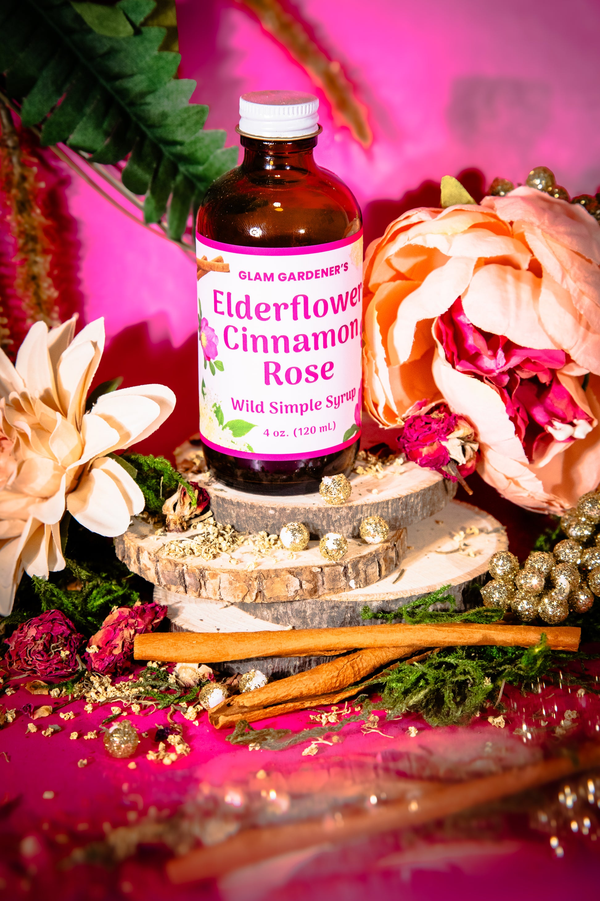 A bottle of Glam Gardeners Elderflower Cinnamon Rose Wild Simple Syrup displayed on a rustic wood slice pedestal, surrounded by cinnamon sticks, dried flowers, and greenery against a vibrant pink background.