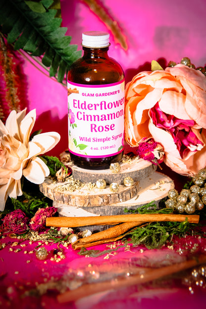 A bottle of Glam Gardeners Elderflower Cinnamon Rose Wild Simple Syrup displayed on a rustic wood slice pedestal, surrounded by cinnamon sticks, dried flowers, and greenery against a vibrant pink background.