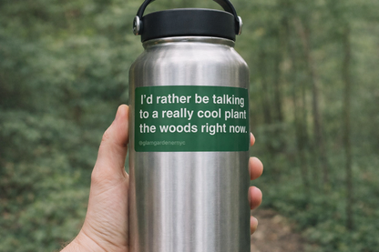 A sticker made by Glam Gardener NYC that says “I’d rather be talking to a really cool plant in the woods right now.” The sticker is hunter green, with white text, is rectangular, and features rounded corners. The image shows the sticker on a water bottle with a woodsy background.