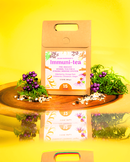 A kraft paper tea box labeled 'Immuni-tea' stands on a wooden tray against a bright yellow background. The box has a floral label and is styled with purple berries, green leaves, and loose tea ingredients, with a mirrored reflection below. By Glam Gardener with Elderberry and Ginger.