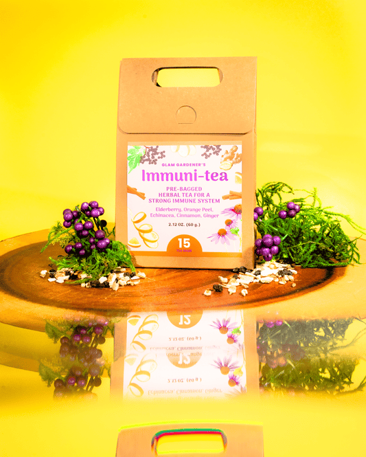 A kraft paper tea box labeled 'Immuni-tea' stands on a wooden tray against a bright yellow background. The box has a floral label and is styled with purple berries, green leaves, and loose tea ingredients, with a mirrored reflection below. By Glam Gardener with Elderberry and Ginger.