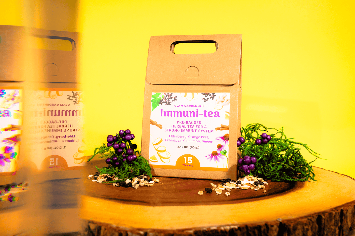 A kraft paper bag of Immuni-tea herbal tea by Glam Gardener stands on a wooden slice with small purple berries and greenery, against a bright yellow background. A second tea bag is blurred in the left foreground. The ingredients have Elderberry, Orange Peel, Echinacea, Cinnamon, Ginger.