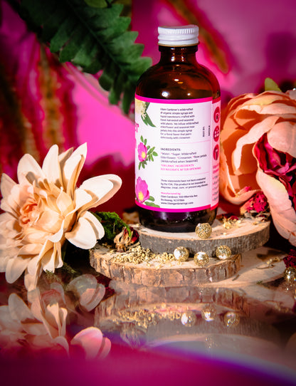 An amber glass bottle of herbal liquid with a pink label and white cap, styled on a wooden slice pedestal with dried herbs, gold beads, and large flowers against a vivid pink background.