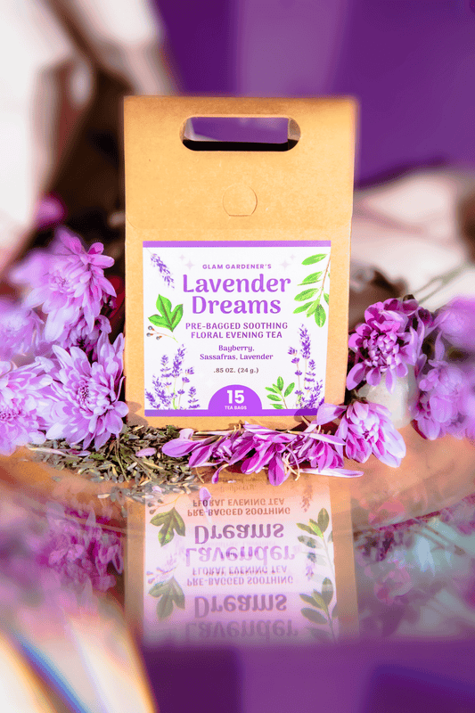 A kraft paper stand bag labeled 'Lavender Dreams' surrounded by purple flowers on a reflective surface with a purple and warm-toned blurred background. By Glam Gardener with Bayberry Sassafras, and Lavender