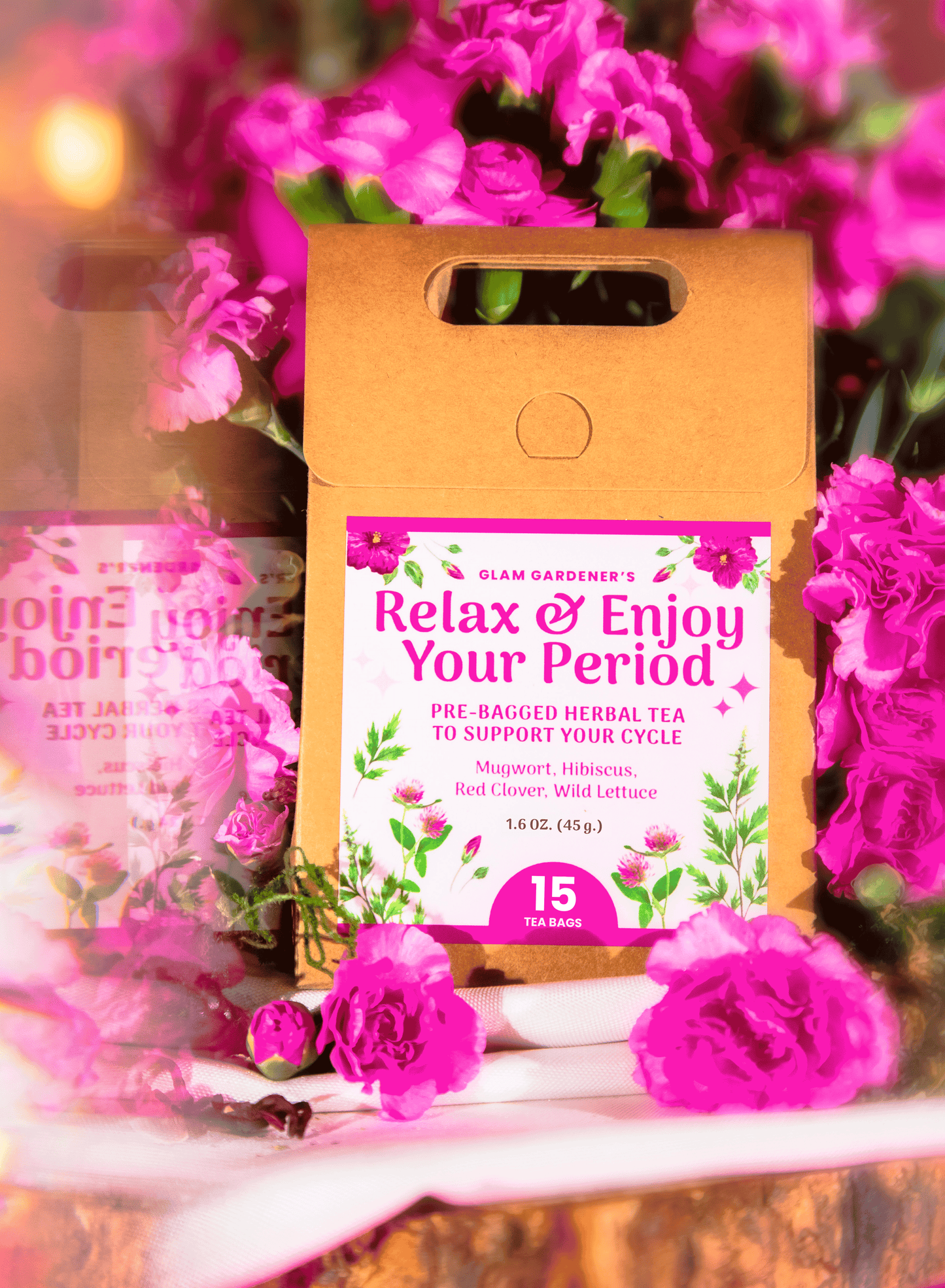 A kraft paper tea box labeled 'Relax & Enjoy Your Period' by Glam Gardener's, surrounded by bright pink flowers on a white surface with a blurred floral background.