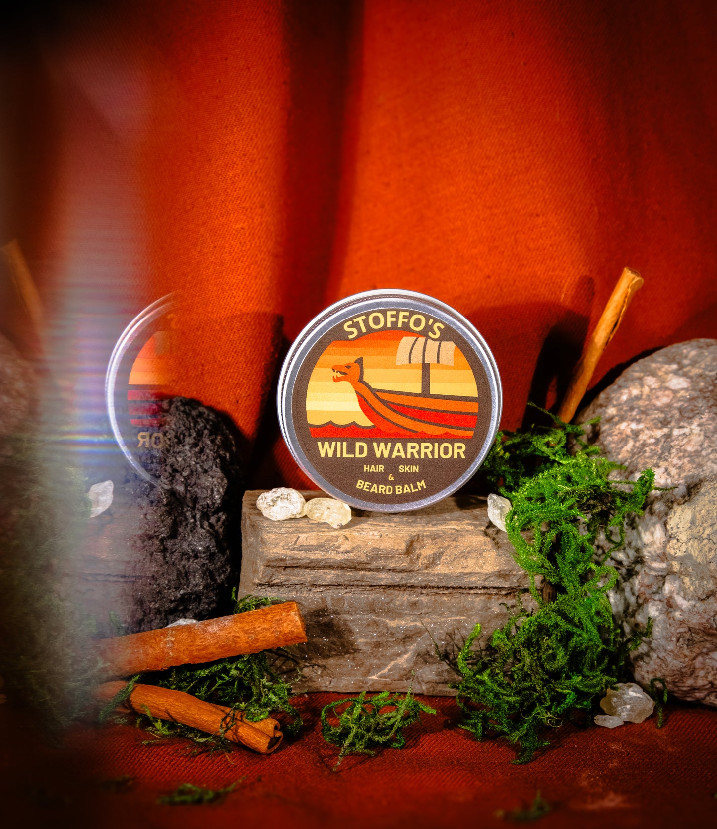 A round tin of Stiffo's Wild Warrior Beard Balm is displayed on a rustic wooden block surrounded by moss, rocks, and cinnamon sticks against a warm orange background.