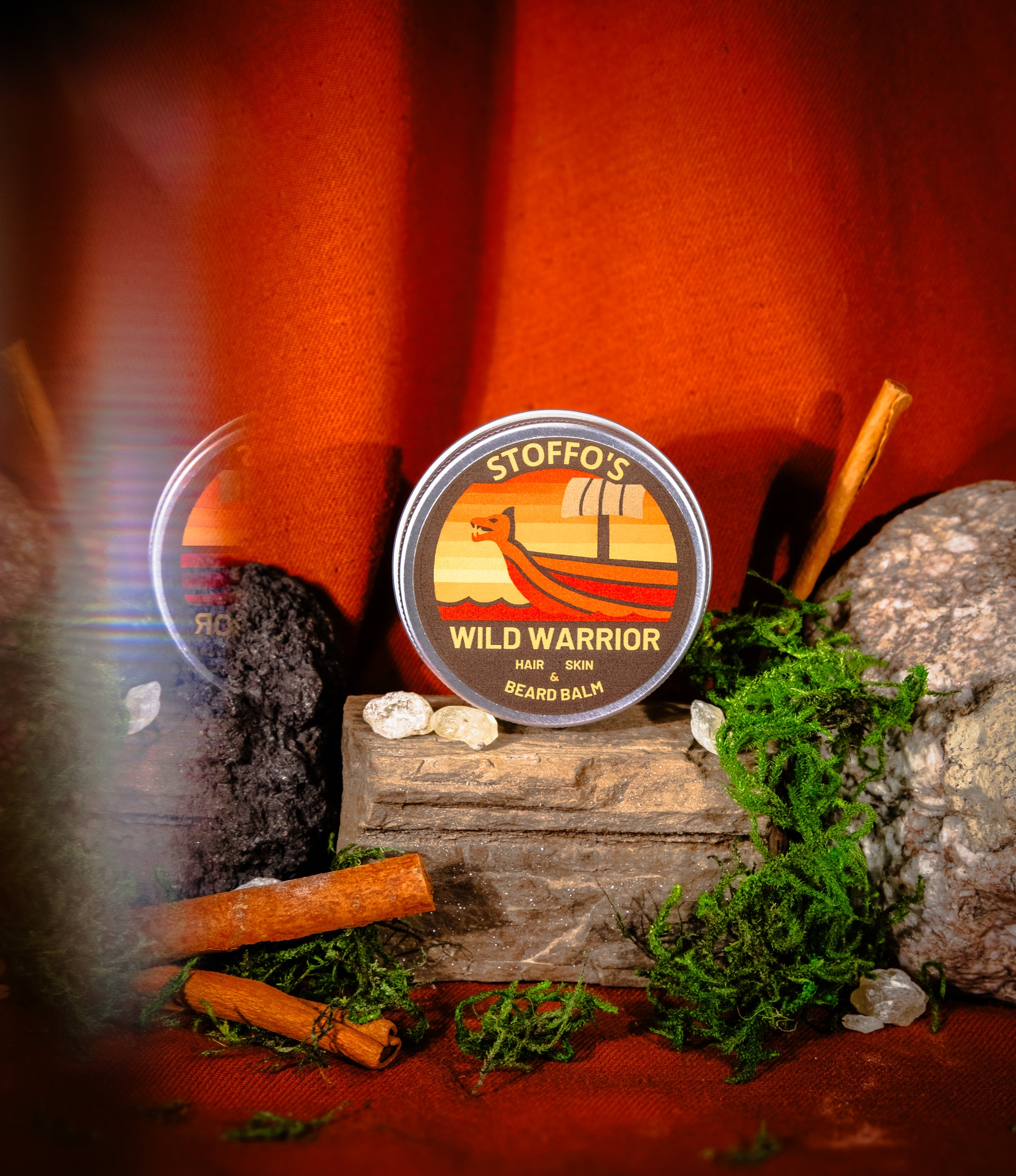 A round tin of Stiffo's Wild Warrior Beard Balm is displayed on a rustic wooden block surrounded by moss, rocks, and cinnamon sticks against a warm orange background.