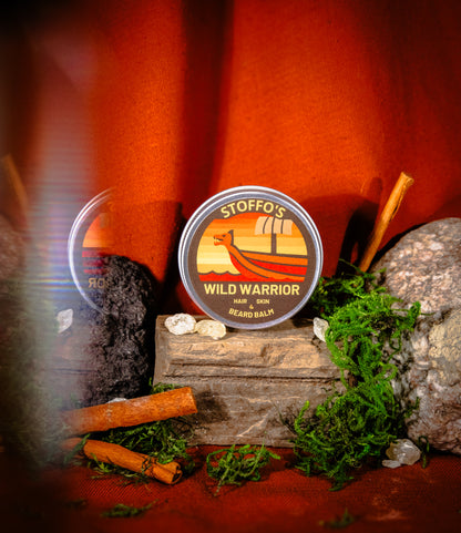 A round tin of Stiffo's Wild Warrior Beard Balm is displayed on a rustic wooden block surrounded by moss, rocks, and cinnamon sticks against a warm orange background.